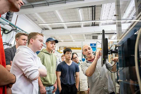 Foto eines Dozenten, der zwischen zwei Studierenden steht und ihnen ein Bauteil erklärt, das er in der Hand hält. 