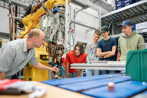 Foto eines Dozenten, der zwischen zwei Studierenden steht und ihnen ein Bauteil erklärt, das er in der Hand hält.
