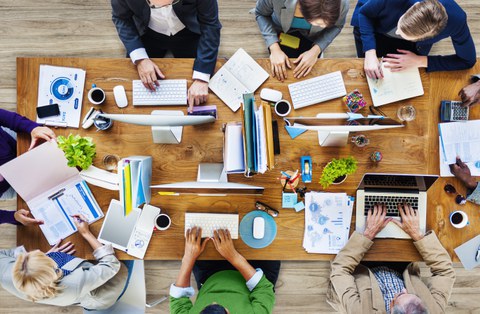 Das Foto zeigt einen großen Tisch aus der Vogelperspektive. Auf dem Tisch stehen Computer, Laptops und weitere Büromaterialien. Um den Tisch herum sitzen mehrer Personen und arbeiten an den Computern.