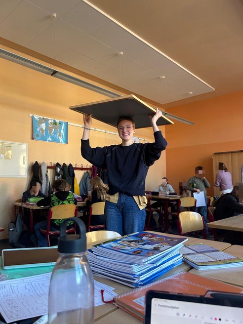 Eine Studentin trägt auf dem Kopf eine große Tafel und lächelt in die Kamera. Sie ist in einem Klassenraum mit Kindern.