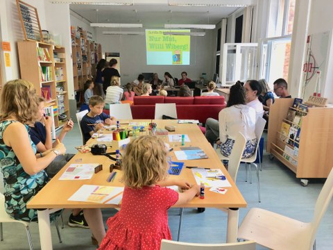In der Lern- und Forschungswerkstatt sitzen Kinder und Erwachsene, die Bilder zum Thema Schule gestalten oder sich ein Bilderbuchkino zum Bilderbuch 'Willi Wiberg!" ansehen.