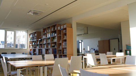 mehrteiliger Raum mit Gruppentischen und Stühlen und einem Regal