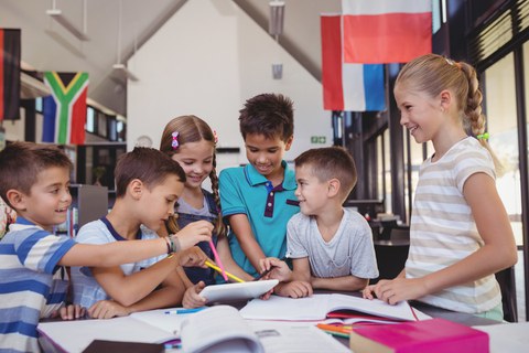 Auf dem Foto sind Kinder um einen Tisch herum versammelt. Auf dem Tisch liegen viele Unterlagen und Stifte. Ein Kind hält ein Tablet, andere schauen hinein und zeigen darauf.