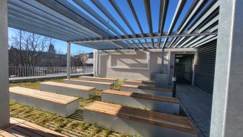 Abgebildet sind wetterbeständige Tische und Bänke für ca. 30 Personen sowie eine Projektionstafel im Freien auf der Wiese hinter dem Fakultätsgebäude. 