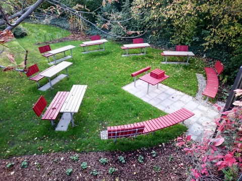 Abgebildet sind wetterbeständige Tische und Bänke für ca. 30 Personen sowie eine Projektionstafel im Freien auf der Wiese hinter dem Fakultätsgebäude. 