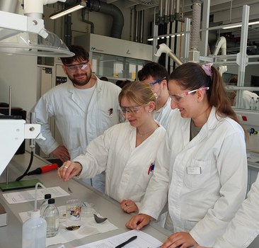 Es stehen zwei Frauen und zwei Männer vor einem Labor Tisch und geben eine Lösung in Wasser.