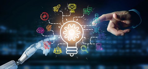 Foto einer menschlichen Hand im oberen rechten Bildrand und einer Roboterhand im unteren linken Bildrand. Beide Händen zeigen mit dem Zeigefinger auf eine visualisierte Darstellung einer Glühbirne mit verschiedenen Icons, die um diese herum angeordnet sin