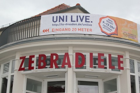 Das Foto zeigt die obere Hälfte einen weißen Gebäudes. Darauf steht mit roten Großbuchstaben das Wort "Zebradiele" geschrieben. Oberhalb davon erkennt man ein Geländer. An diesem ist ein Plakat befestigt. Es trägt die Aufschrift: "Uni Live. 
