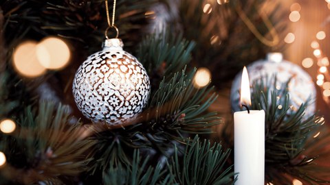 Das Foto zeigt eine Nahaufnahme eines geschmückten Weihnachtsbaumes mit einer brennenden Kerze und zwei Kugeln.