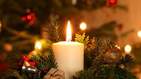 Das Fto zeigt eine weiße, brennende Stumpenkerze. Sie steckt in einem Weihnachtsgesteck aus Tannengrün-Zweigen und Dekoration wie Christbaumkugeln und kleinen Geschenken. Unscharf im Hintergrund sieht man einen geschmückten Tannenbaum.