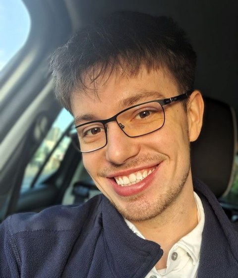 Pascal, ein junger Mann mit dunklen Haaren und Brille, lächelt strahlend in die Kamera.