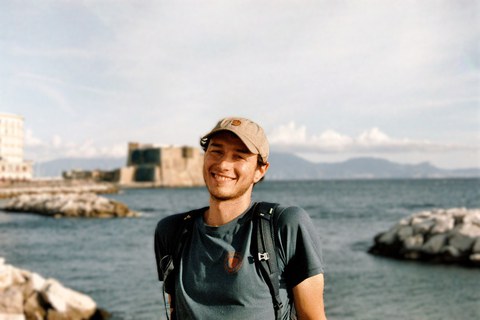Paul, ein junger Mann mit dunklen Haaren, schaut freundlich in die Kamera. Hinter ihm ist das Meer zu sehen.