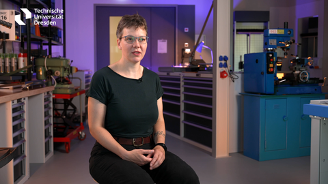 Frau Andreas, eine junge Frau mit kurzen Haaren und dunkler Kleidung, sitzt auf einem Stuhl. 