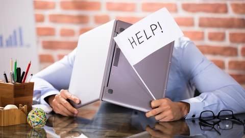 Foto eines Mannes am Schreibtisch. Er hält eine Fahne mit der Aufschrift "Help" und einen aufgeklappten Laptop in den Händen. Hinter diesem versteckt er seinen Kopf.