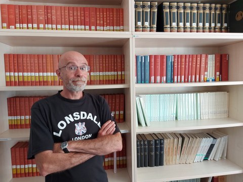 Florent Cygler steht vor einem Bücherregal in der Bibliothek der FOVOG. Er trägt ein schwarzes T-Shirt und verschränkt die Arme.