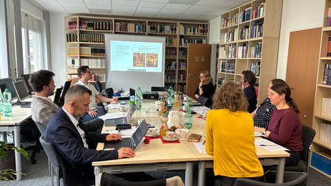 Blick in die Bibliothek der FOVOG. Viele Menschen sitzen an einem Tisch, im Hintergrund ist eine Präsentation zu sehen. 