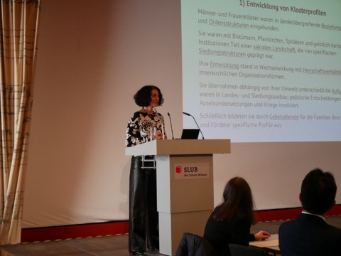 Christina Lutter steht hinter einem Podium, auf dem das SLUB-Logo zu sehen ist