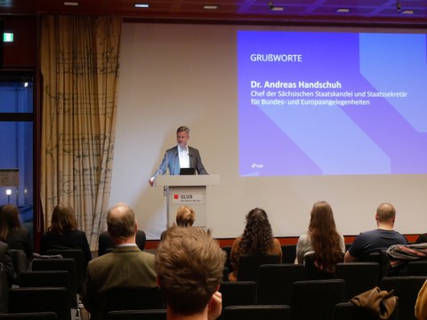 Andreas Handschuh stands behind a lectern. On his left is a presentation stating "Grußworte. Dr. Andreas Handschu. Chef der Sächsischen Staatskanzlei und Staatssekretär für Bundes- und Europaangelegenheiten".