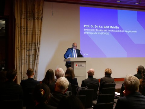Gert Melville steht hinter dem Podium. Zu seiner Linken ist eine Präsentation im TUD-Design zu sehen, auf der steht: "Prof. Dr. Dr. h.c. Gert Melville. Emeritierter Direktor der Forschugnsstelle für Vergleichende Ordensgeschichte (FOVOG)"