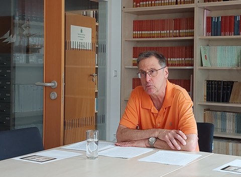 Auf dem Bild ist Bruce Brasington zu sehen. Er trägt ein orangefarbenes Poloshirt und sitzt mit übereinandergelegten Armen an einem Tisch. Er schaut auf an der Kamera vorbei und spricht. Seine Notizen lieg die auf dem Tisch vor ihm. Vor diesen Notizen steht ein Wasserglas auf dem Tisch. In der rechten unteren Bildecke liegen weitere Notizen. Im Hintergrund ist ein Bücherregal sowie zwei Zimmertüren zu sehen. 