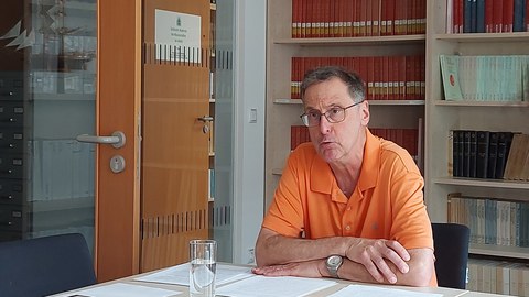 Auf dem Bild ist Bruce Brasington zu sehen. Er trägt ein orangefarbenes Poloshirt und sitzt mit übereinandergelegten Armen an einem Tisch. Er schaut auf an der Kamera vorbei und spricht. Seine Notizen lieg die auf dem Tisch vor ihm. Vor diesen Notizen steht ein Wasserglas auf dem Tisch. In der rechten unteren Bildecke liegen weitere Notizen. Im Hintergrund ist ein Bücherregal sowie zwei Zimmertüren zu sehen. 