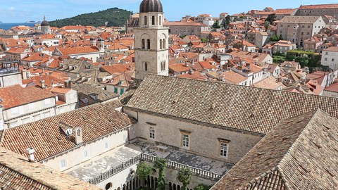 Ansicht des franziskanischen Klosters in Dubrovnik in Kroatien. Im Vordergrund das Kloster mit Blick von oben in den Innenhof mit Kreuzgang, im Hintergrund viele Häuser, dahinter ein Hügel und das Meer.