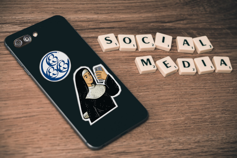 Auf dem Bild ist ein Smartphone zu sehen, das auf einer Holzoberfläche liegt. Auf der Rückseite des Smartphones sind zwei Sticker angebracht: das FOVOG-Logo und eine gezeichnete Nonne mit Smartphone. Neben dem Smartphone liegen Scrabble-Buchstaben, die die Worte "Social Media" formen.