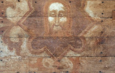 Mittelalterliche Tafelmalerei auf waagerechten Holzplanken, die ein bärtiges Gesicht mit großen Augen und Heiligenschein zeigt. Der Kopf ist von einer rötlich-braunen, sternen- oder blütenförmigen Umrahmung umgeben. Die Farben wirken stark verblasst, und auf dem Holz sind einzelne Nagelköpfe sowie Risse in der Oberfläche zu erkennen.