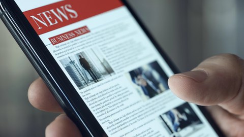 Eine Hand hält ein Smartphone mit geöffneter Nachrichten-App, die eine Überschrift 'NEWS' und mehrere Textabschnitte mit Bildern zeigt.