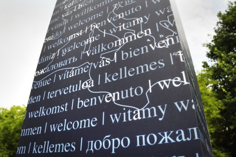Foto einer Textsäule, auf der sich der Gruß Willkommen in verschiedenen Sprachen befindet