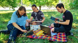 Foto zeigt drei Studierende auf einer Picknickdecke auf einer Wiese. Sie schauen in Geschichtslehrbücher oder Tablets. Die Studierenden lachen.