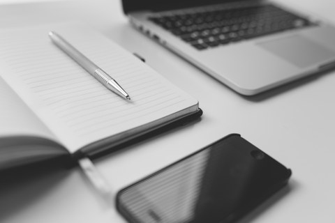 Auf dem Bild sind ein aufgeklappter Notizblock mit Stift, ein Notebook und ein Smartphone zu sehen.