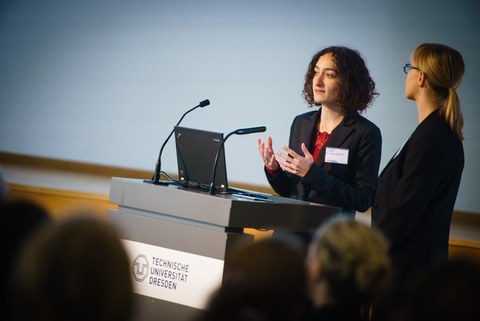 Zu sehen sind zwei Personen an einem Pult, die etwas vortragen.