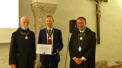 Ein Mann im Anzug, flankiert von zwei Ordensbrüdern, hält eine Ernennungsurkunde in die Kamera.
