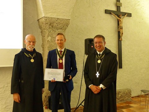 Ein Mann im Anzug, flankiert von zwei Ordensbrüdern, hält eine Ernennungsurkunde in die Kamera.