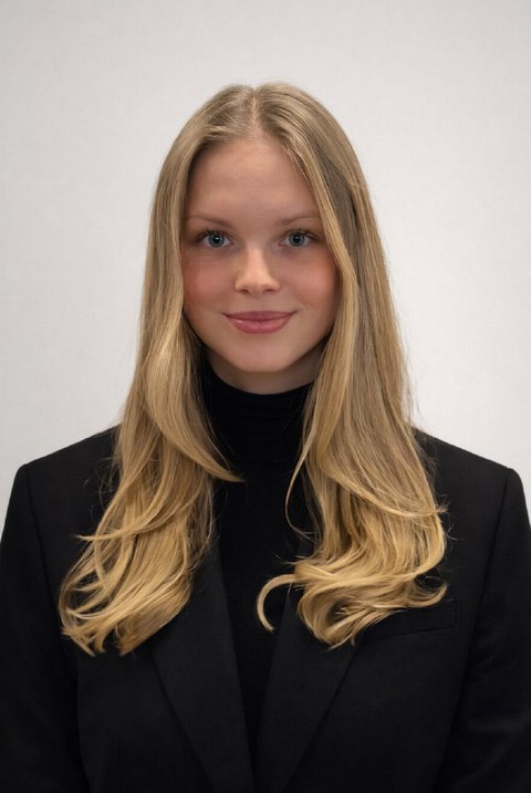 Eine junge Frau in schwarzem Blazer steht lächelnd vor einer weißen Wand.