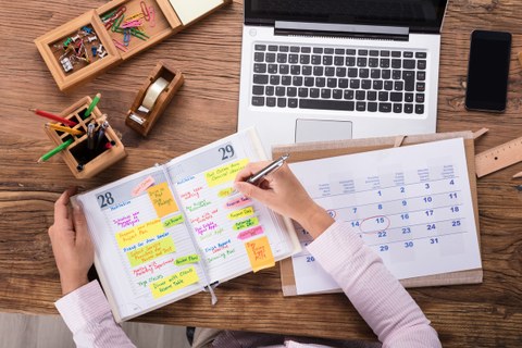 Das Foto zeigt einen aufgeschlagenen Terminkalender auf einem Schreibtisch. Eine Frau notiert etwas in dem Kalender. Auf dem Tisch stehen außerdem ein Laptop und weitere Büromaterialien.