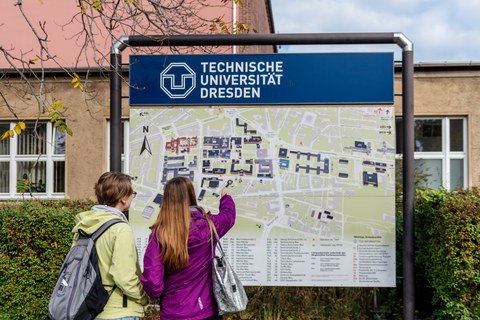 Das Foto zeigt eine Informationstafel. Auf dieser ist die Lage der einzelnen Gebäude auf dem Campus der TU Dresden dargestellt. Vor der Tafel stehen zwei Studentinnen. Eine zeigt mit ihrem Finger auf ein Gebäude auf der Tafel.