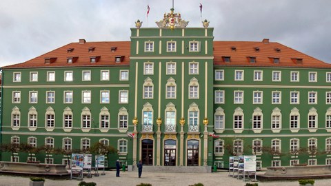 Zu sehen ist die Hauptfront des Rathauses von Stettin mit dem Wappen der Stadt auf dem Giebel. Auf dem Platz sind zwei Zierbrunnen und kleine Bäume.