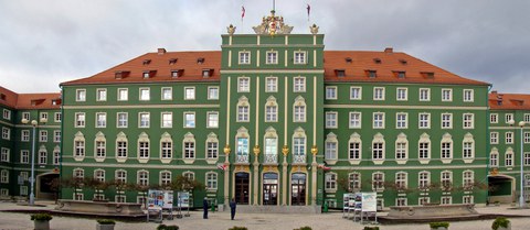 Zu sehen ist die Hauptfront des Rathauses von Stettin mit dem Wappen der Stadt auf dem Giebel. Auf dem Platz sind zwei Zierbrunnen und kleine Bäume.