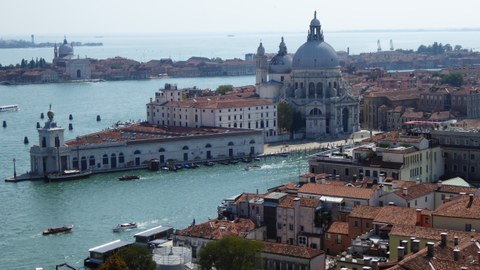 Küste von Venedig, man sieht das Wasser, darauf Boote, im Hintergrund eine Kirche und viele Dächer