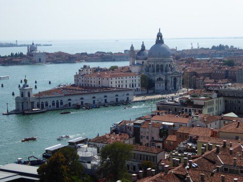 Küste von Venedig, man sieht das Wasser, darauf Boote, im Hintergrund eine Kirche und viele Dächer