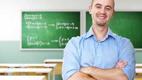 Das Foto zeigt einen Lehrer in seinem leeren Klassenraum. Er hat die Arme vor dem Körper verschränkt und lächelt in die Kamera.