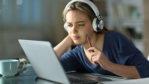 Das Foto zeigt eine junge Frau an einem Tisch. Sie schaut nachdenklich auf ihren Laptop.