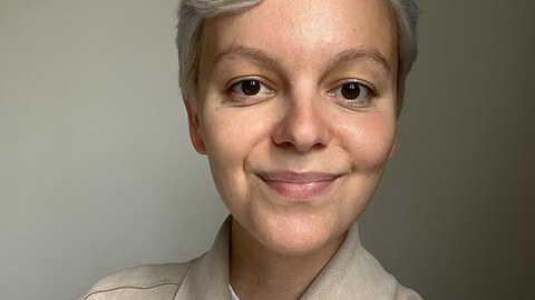 Eine jung und weiblich gelesene Person mit Kurzhaarschnitt lächelt freundlich in die Kamera. 