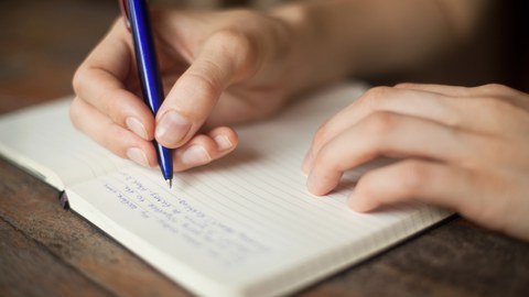 Foto zeigt ein geöffnetes Notizbuch und ein Hand mit einem Stift