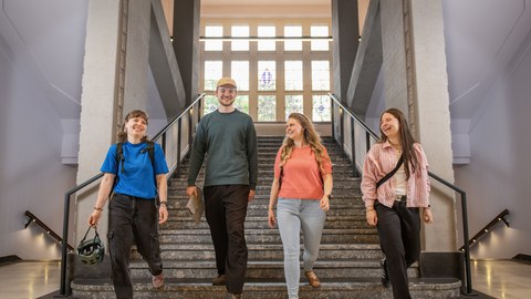 Foto: Vier Studierende, die lachend eine breite Treppe hinuntergehen, im Hintergrund ist ein großes Fenster zu sehen.