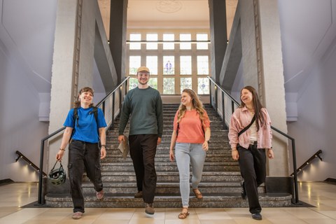 Foto: Vier Studierende, die lachend eine breite Treppe hinuntergehen, im Hintergrund ist ein großes Fenster zu sehen.