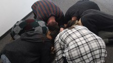 sechs Personen knien mit gesenktem Oberkörper auf dem Boden und bilden einen Kreis