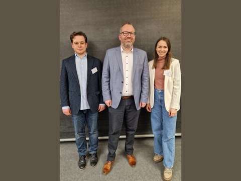 Drei erwachsene Personen stehen nebeneinander vor einer dunklen Wand, frontal zur Kamera. Alle tragen legere bis formelle Kleidung mit Namensschildern; links ein Mann im Sakko, in der Mitte ein Mann im hellblauen Sakko, rechts eine Frau im hellen Blazer.